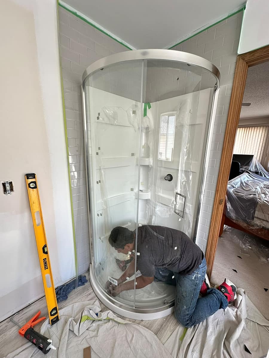 Bathroom Renovation (Tamarack) — installer kneeling to set the shower base tracks with a caulking gun and level nearby