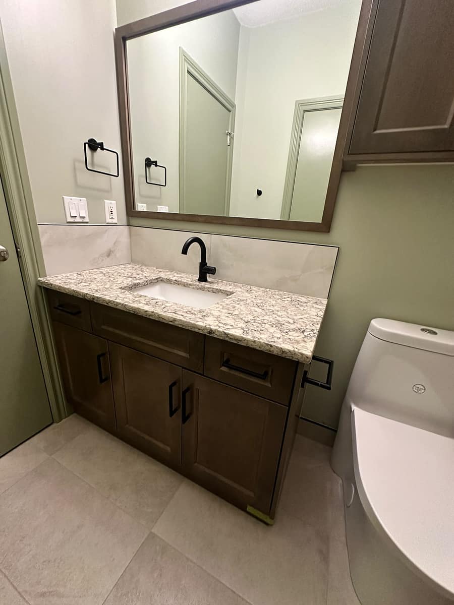 Bungalow Home Renovation — vanity close-up with black faucet, speckled countertop, large mirror and modern toilet against sage green walls