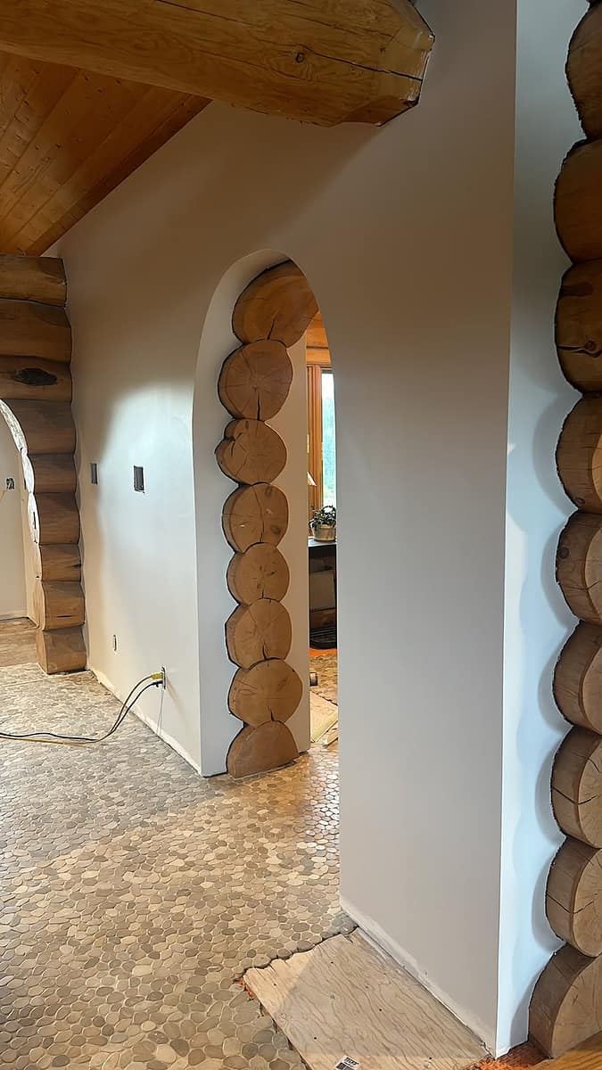 Custom Log Home Interior Renovation — arched doorway cut through the white-painted wall revealing the log ends, pebble-tiled floor transitioning to subfloor