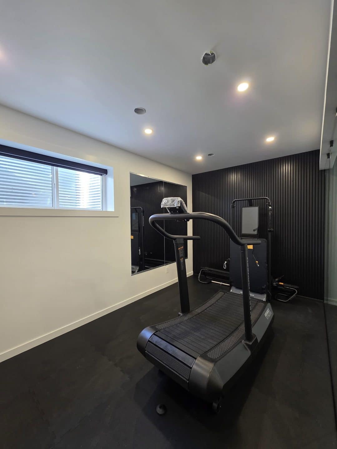 Basement Development — inside the glass gym showing Assault Fitness curved treadmill, digital strength machine and black slat wall