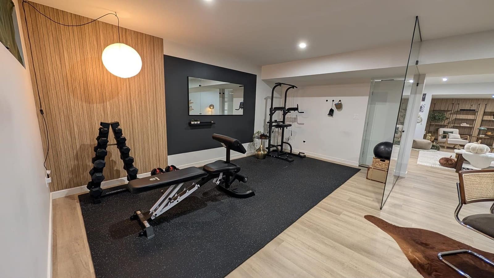 South Edmonton basement home gym with dumbbell rack, adjustable weight bench and power tower on a black rubber floor beside the glass partition